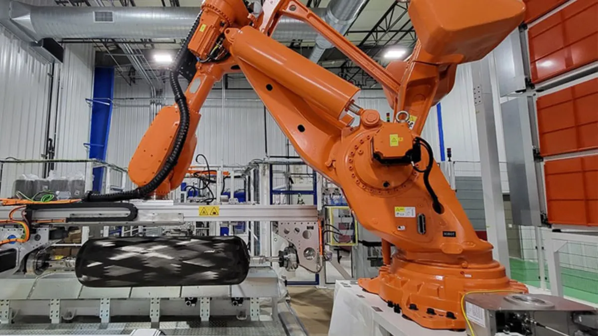 An orange robotic arm working on a black hydrogen storage tank in a white and gray facility.