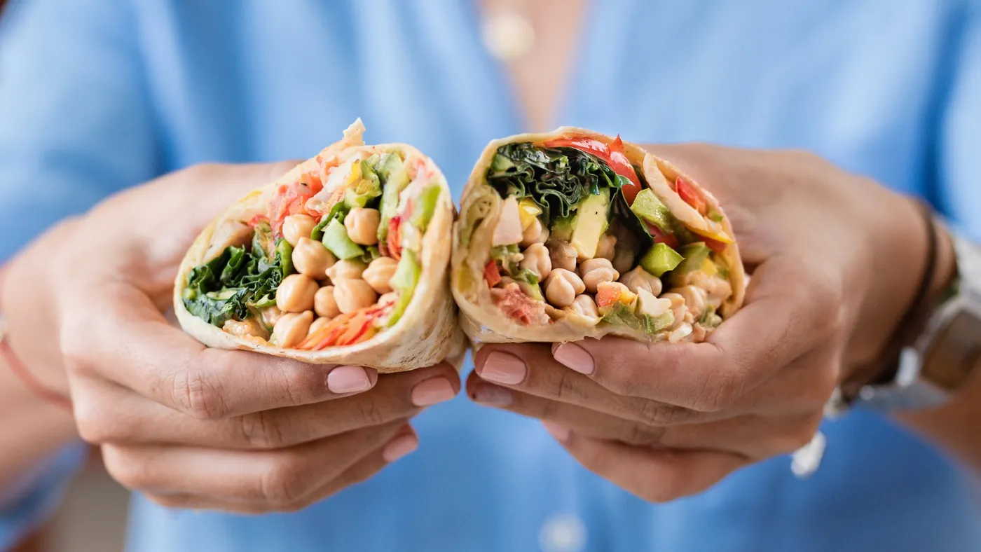Hands holding a colorful, halved wrap filled with chickpeas, leafy greens, tomatoes, and avocado. The background is softly blurred, suggesting a casual setting.