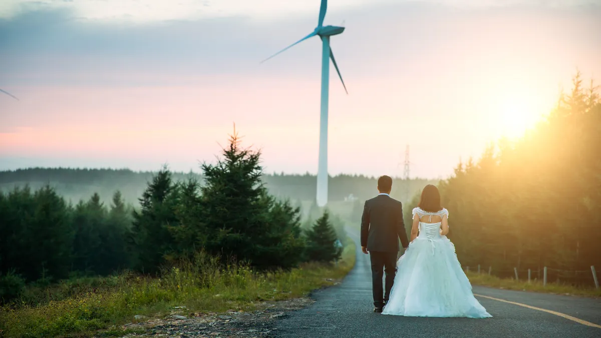 A couple seen from behind in a black suit and a long white dress walk down a country road toward a wind turbine as the sun sets.