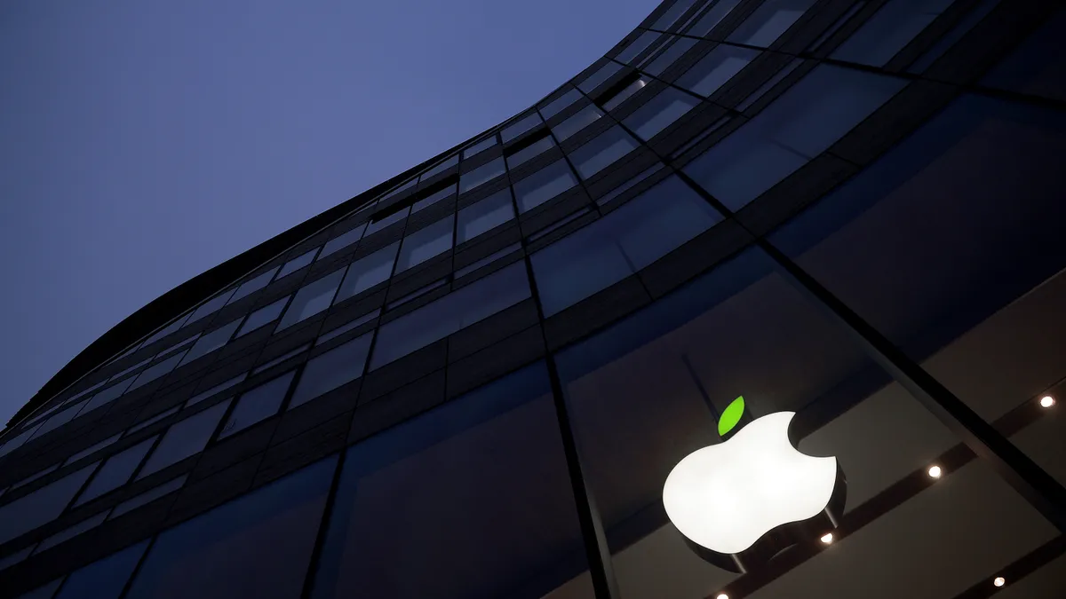 A green leaf on top of a white Apple logo outside a glass building at night.