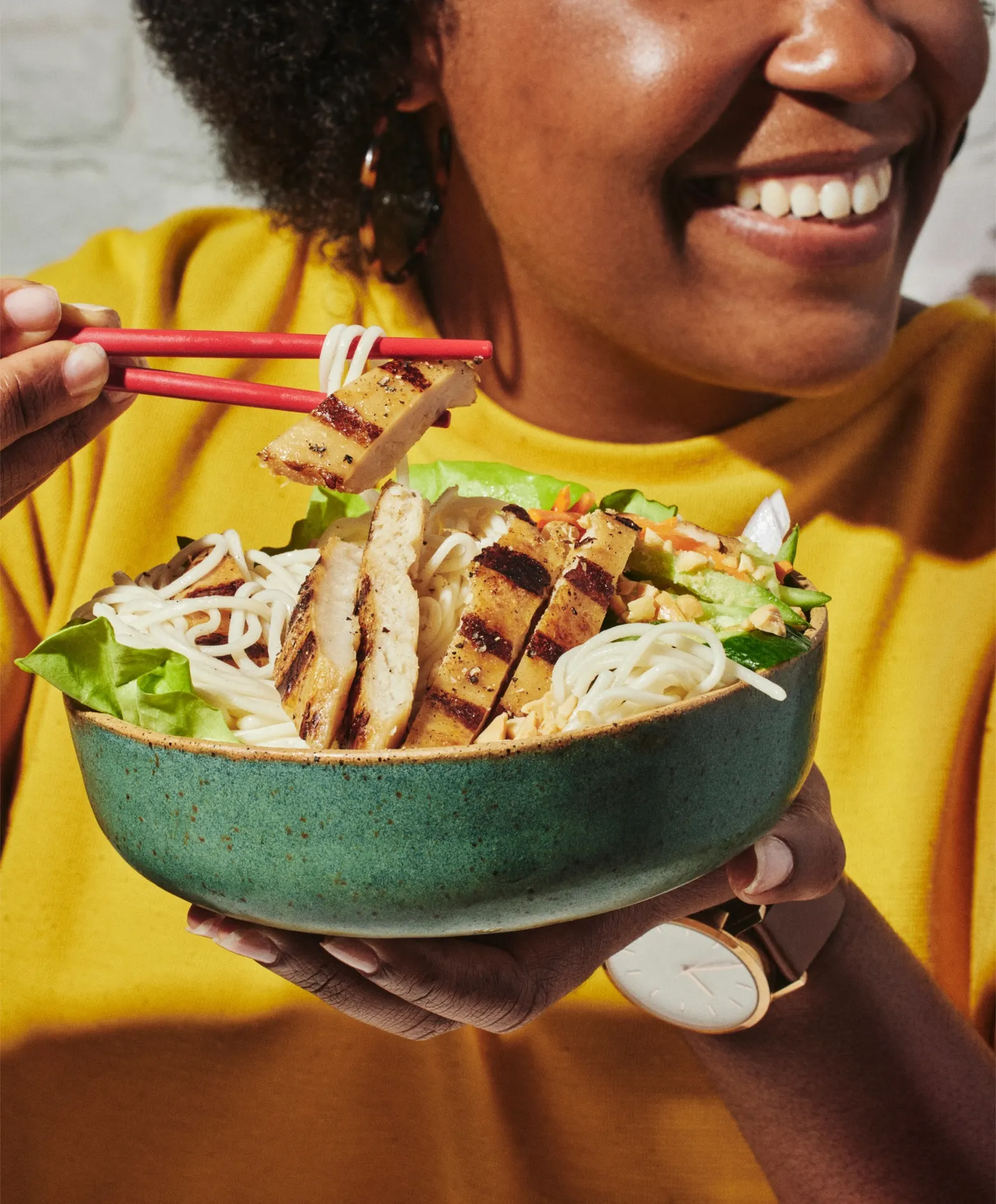 A person in a yellow shirt uses red chopsticks to pick up grilled cultivated chicken and ramen noodles in a green bowl.