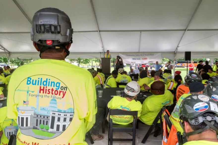 People in bright yellow shirts and hard hats gather under a tent at an event.