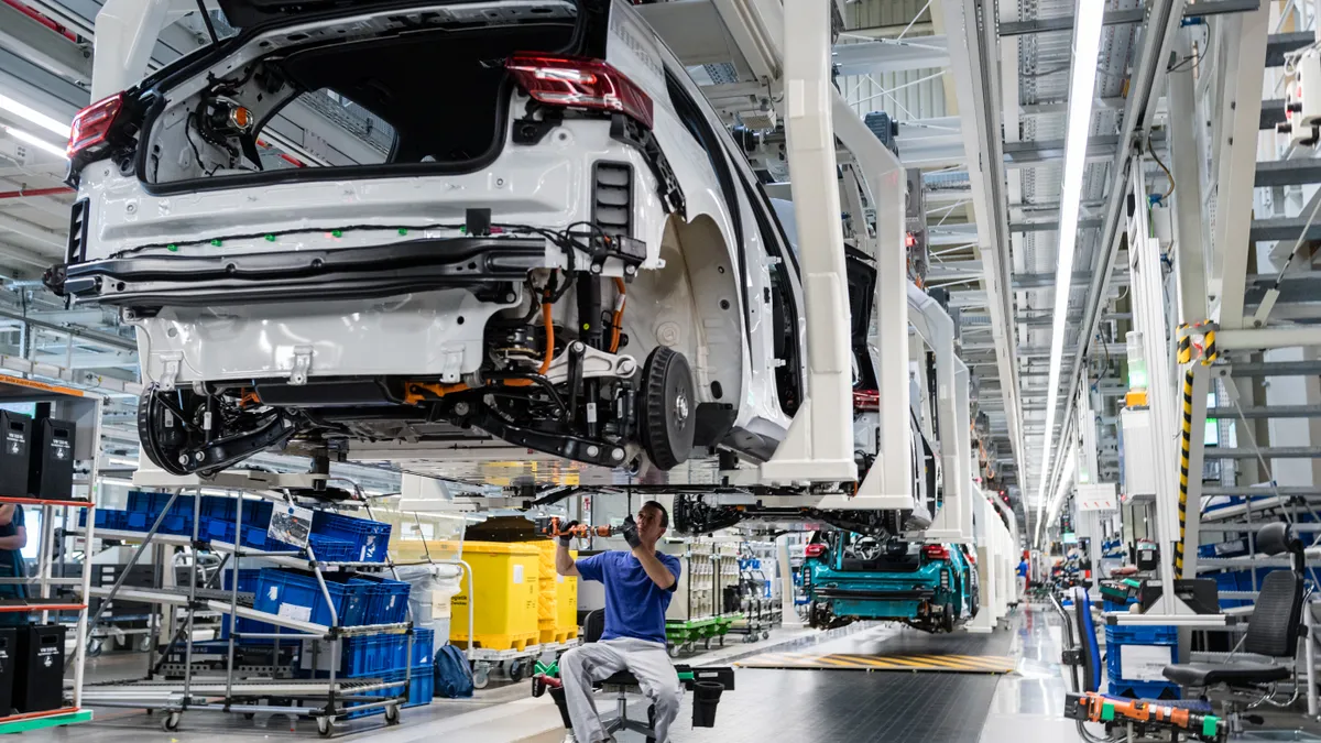 A worker sits below a white, unfinished vehicle and holds a power tool to complete assembling the car.