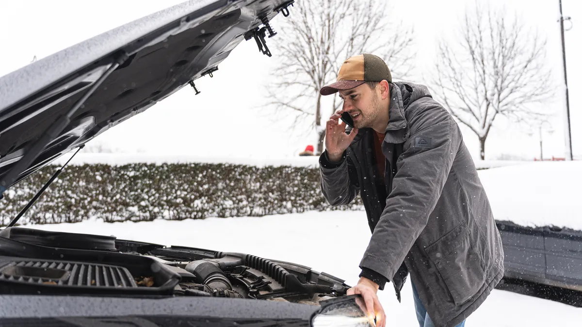 A man, talking on mobile phone while looking at the car engine on the side of a road.