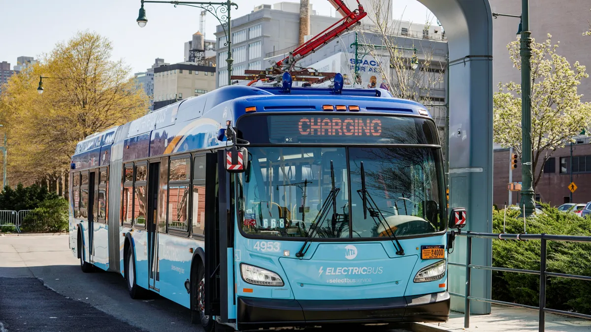 A blue electric New York City transit bus at an overhead charging station.