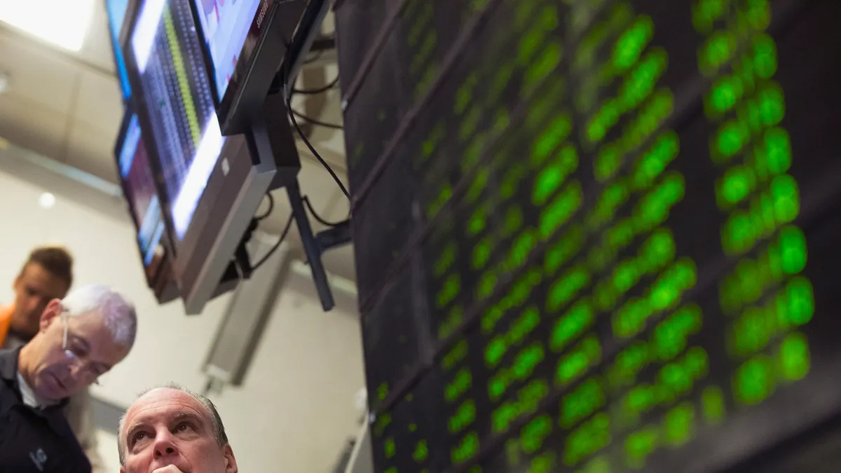 A stock trader with hand on chin in the bottom left looks up at market screens with out of focus TVs showing the S&P 500.