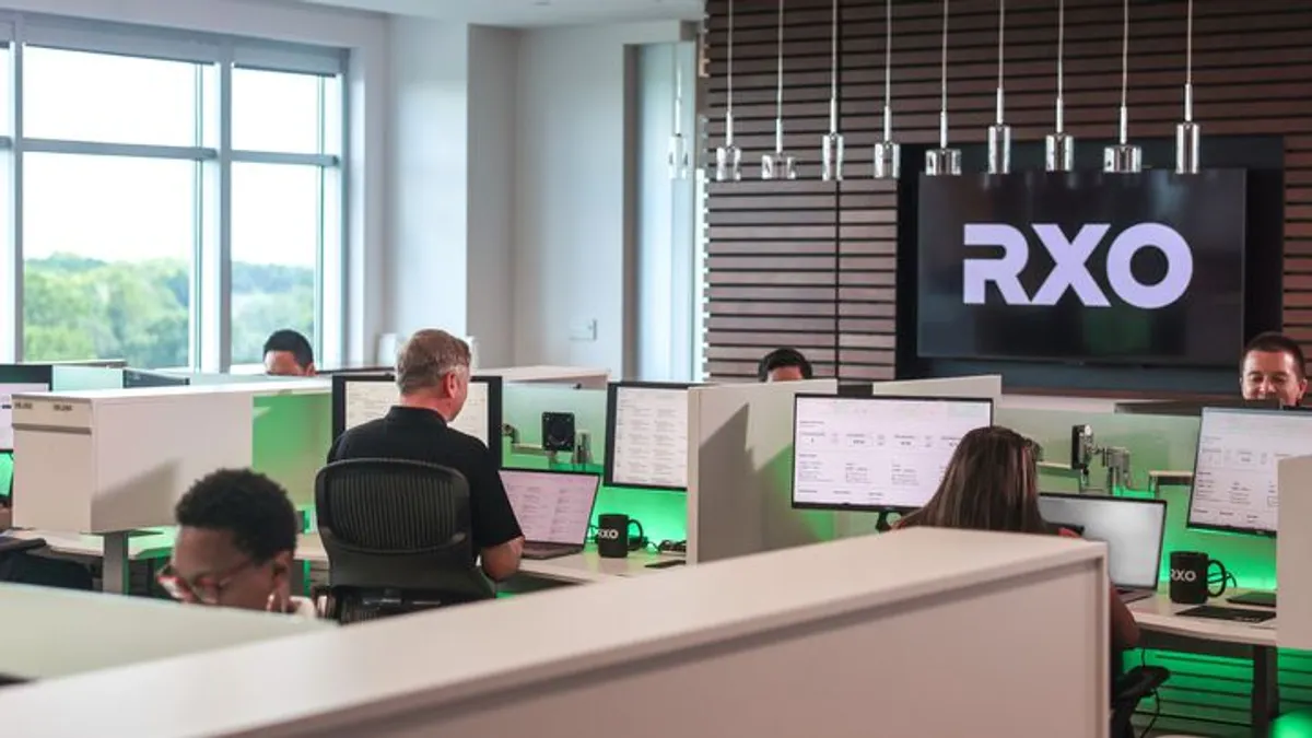 RXO staff work at computers in a modern office with a TV screen showing the company's logo and green lights illuminating desk dividers.