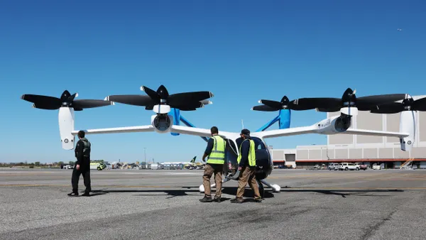 Two men wearing safety vests and another man inspect an electric vertical takeoff and landing aircraft with a large building and a passenger aircraft in the distance.