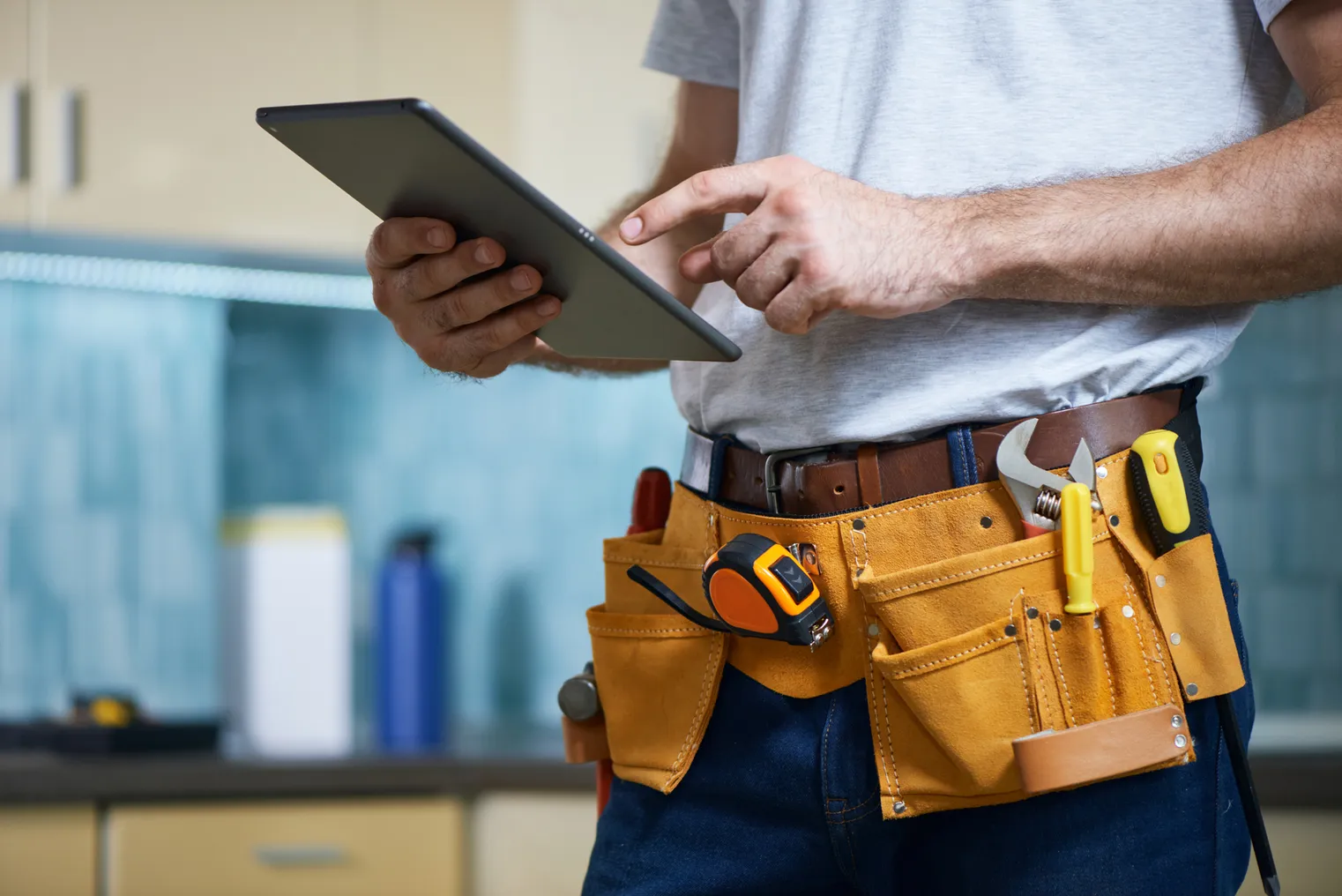 A cropped shot of a young repairman wearing a tool belt with various tools using digital tablet while standing indoors.