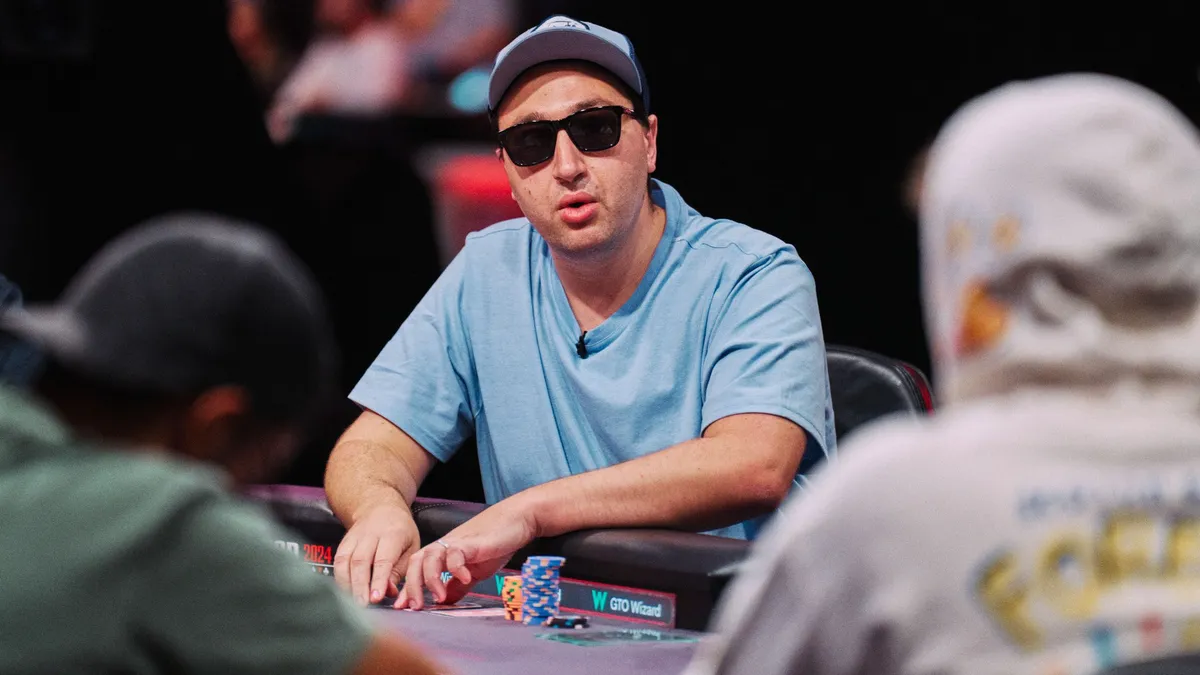 A person wearing a hat and sunglasses sits at a poker table facing two other people.