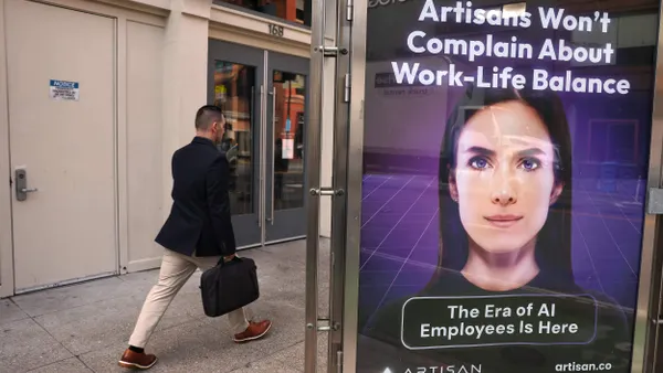 A person carrying a briefcase walks past a sidewalk advertisement featuring a digitally rendered face and the text “Artisans Won’t Complain About Work-Life Balance” and “The Era of AI Employees Is Here.