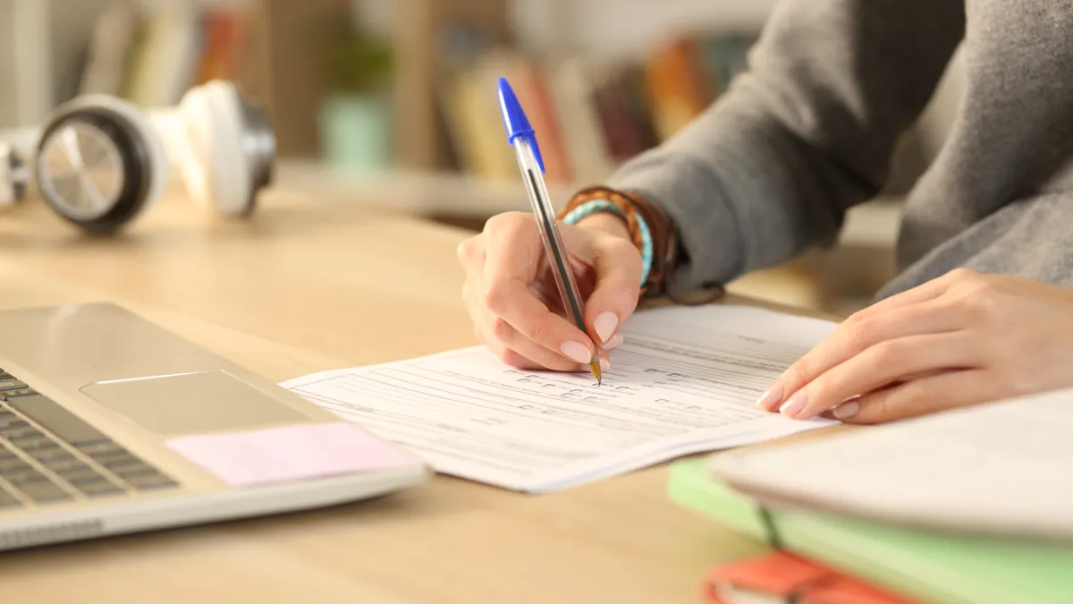 A person writes at a desk that also holds a laptop.