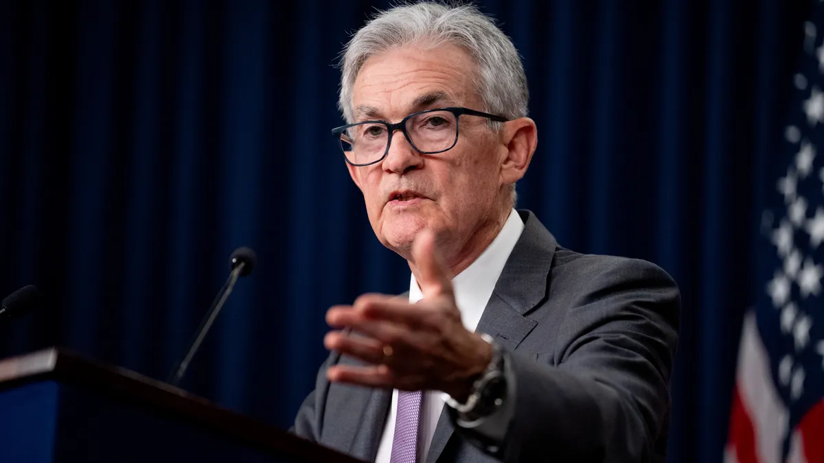 Federal Reserve Chairman Jerome Powell gestures while speaking at a podium with a blue curtain and America flag behind him in the background.