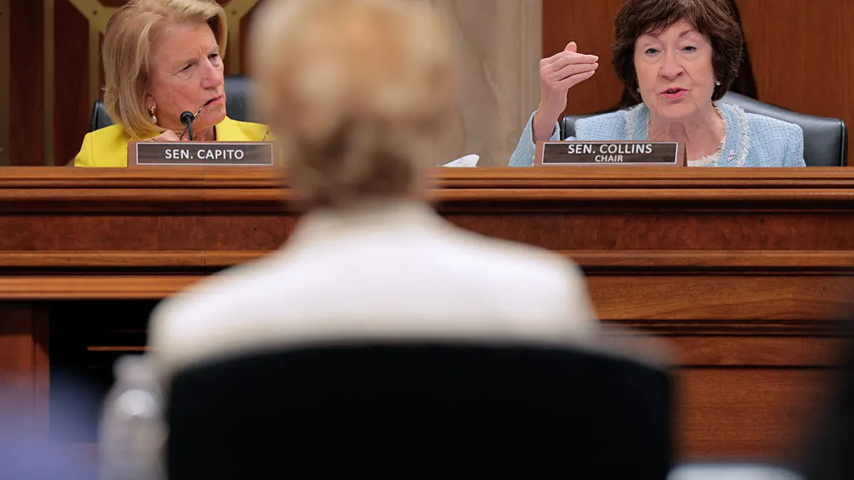 Two people are seated at a dias in a hearing room. They are facing the camera and looking at one person seated at a desk whose back is to the camera.