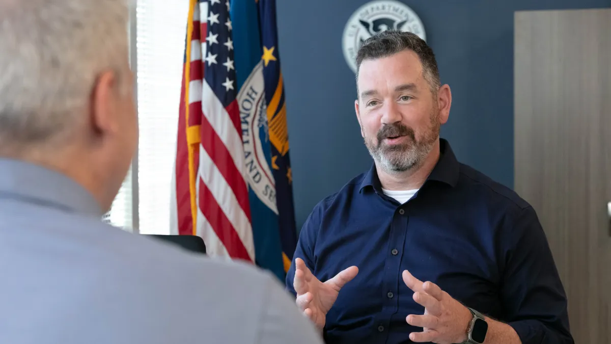 Trent Frazier speaks with someone as a U.S. Department of Homeland Security flag hangs next to an American flag behind Frazier.