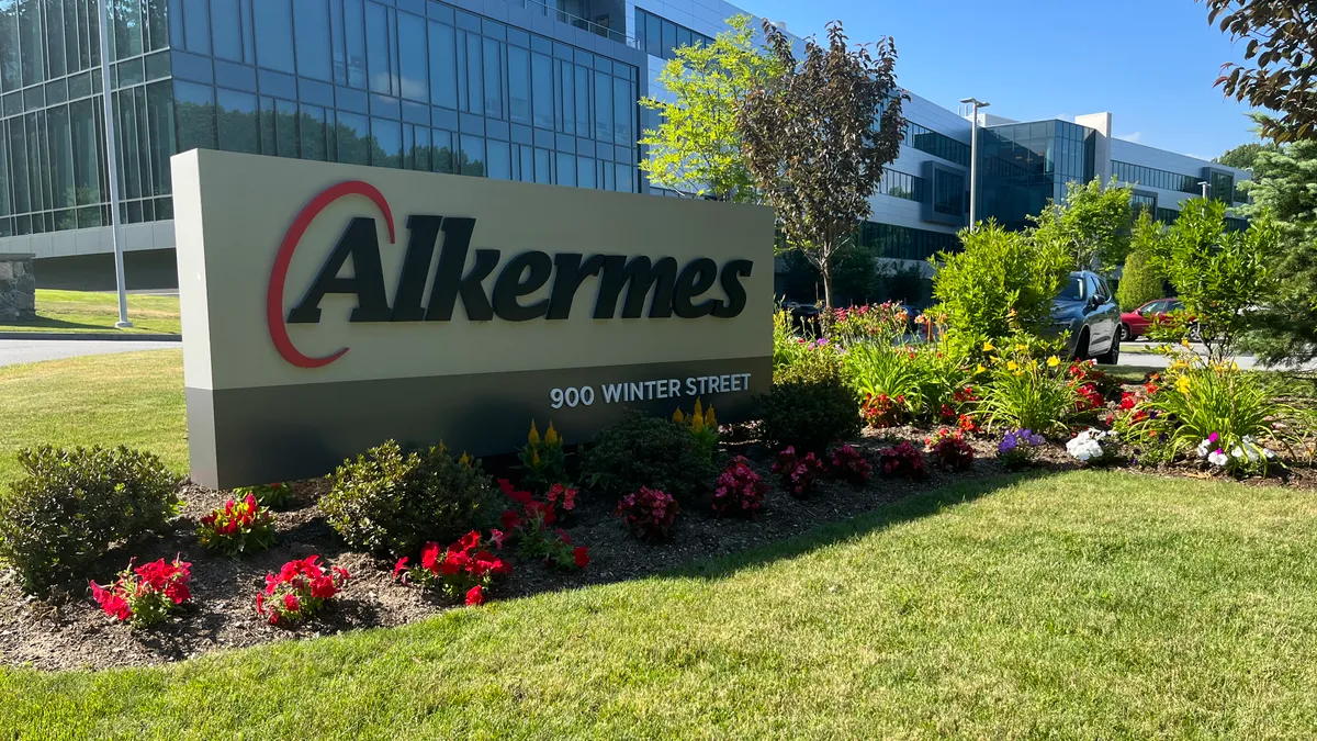 A grey exterior sign with Alkermes' written on it sits in a bed of flowers outside a glass office building.