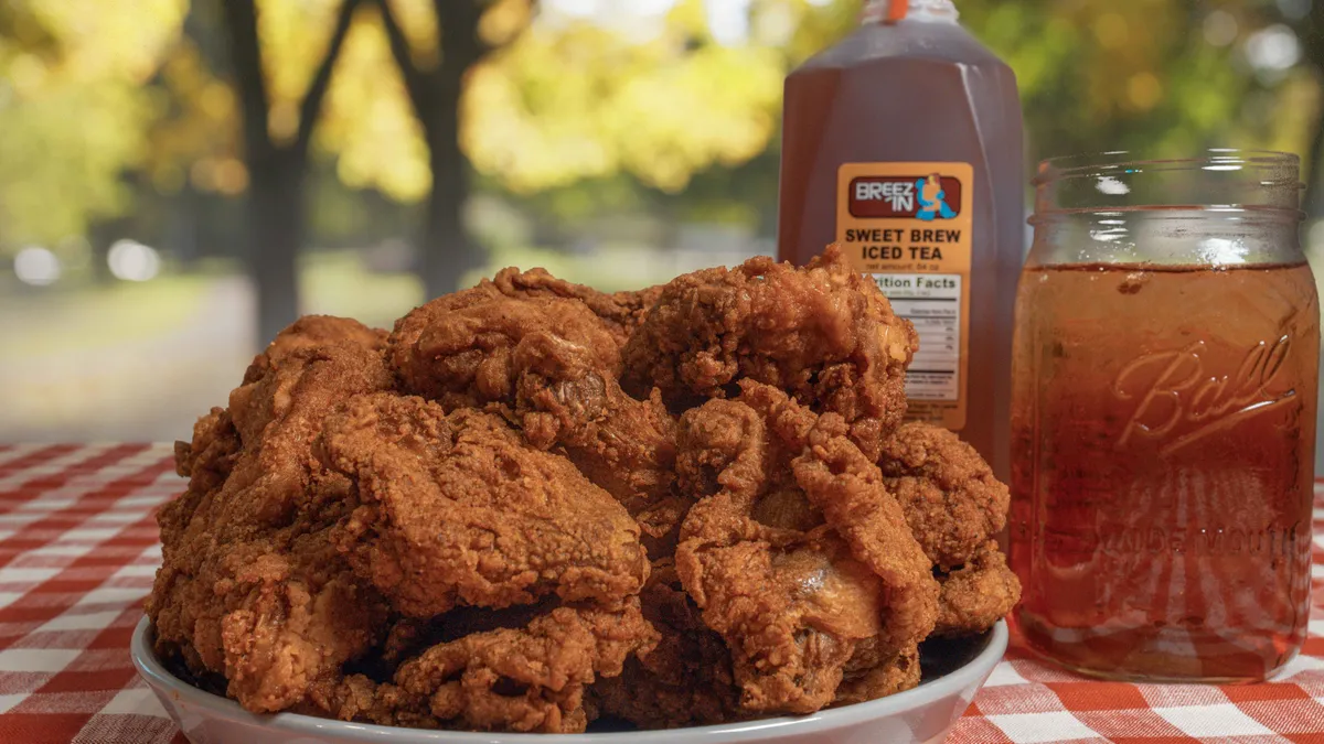 A photo of a place of fried chicken on a table outdoors with a red checkered tablecloth under it and a glass and jug of tea behind it.