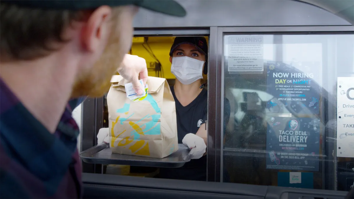 A Taco Bell employee wearing a mask hands a drive-thru customer their order.