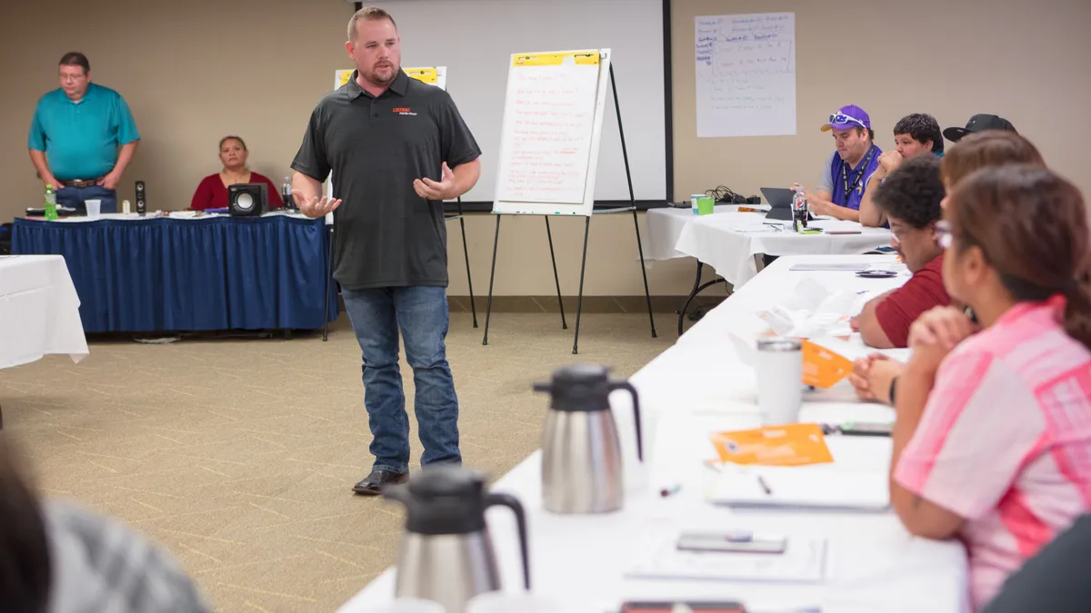 A speaker in a polo stands in front of a room of people taking notes at tables.