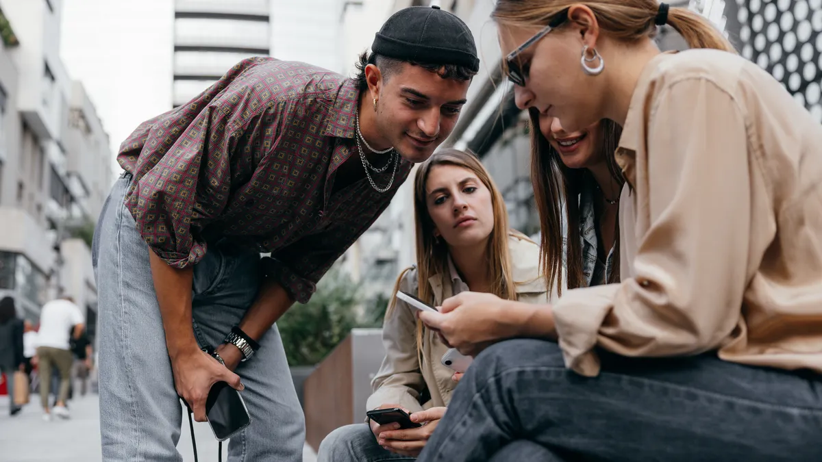 A group of four young people gather around in a semi-circle to look at what's on one of their phones.