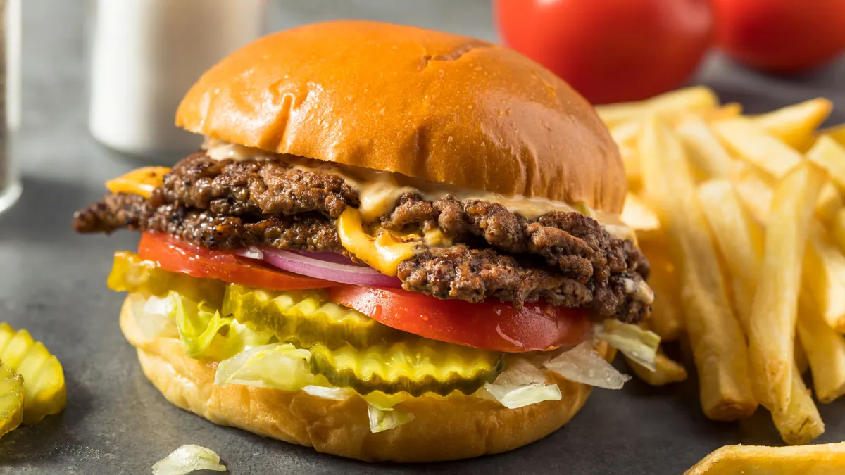 A close up photograph of a cheeseburger with tomatoes, red onions, lettuce and pickles.