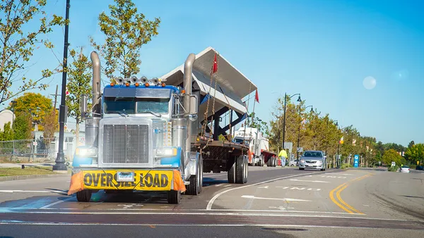 An eighteen wheeler truck at a stop sign with an oversize load on its bumper.