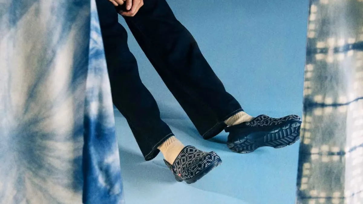 A person sitting amidst a blue background wearing jeans and navy blue denim Levi's Crocs.