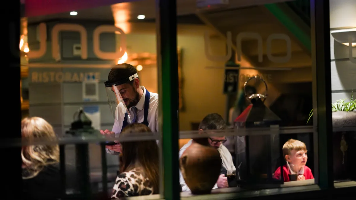 A restaurant worker wearing a clear plastic mask shield speaks with restaurant patrons.