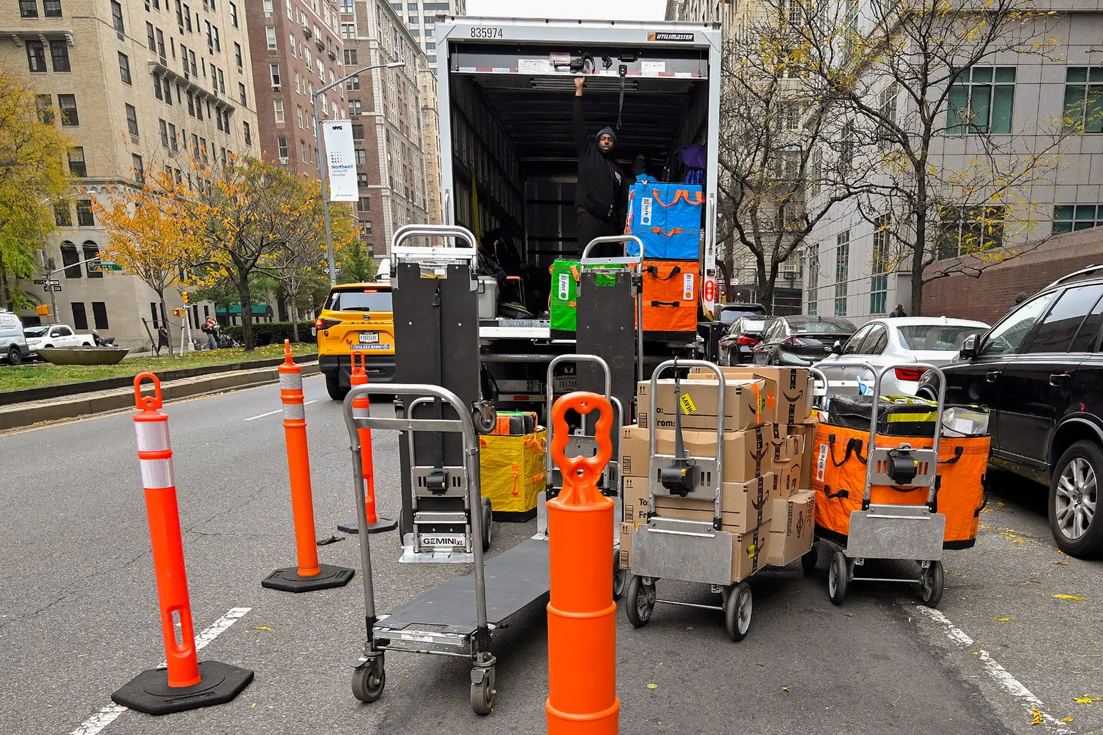 Flat wagons carrying parcel shipments or are empty sit in front of an open truck holding good in the street. The truck is surrounded by orange cones, with cars parked alongside the right side of the vehicle, as well as buildings in the background.