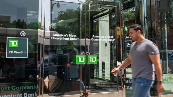 A person walks past the metal and glass doors of a TD Bank branch. The doors read "America's Most Convenient Bank."