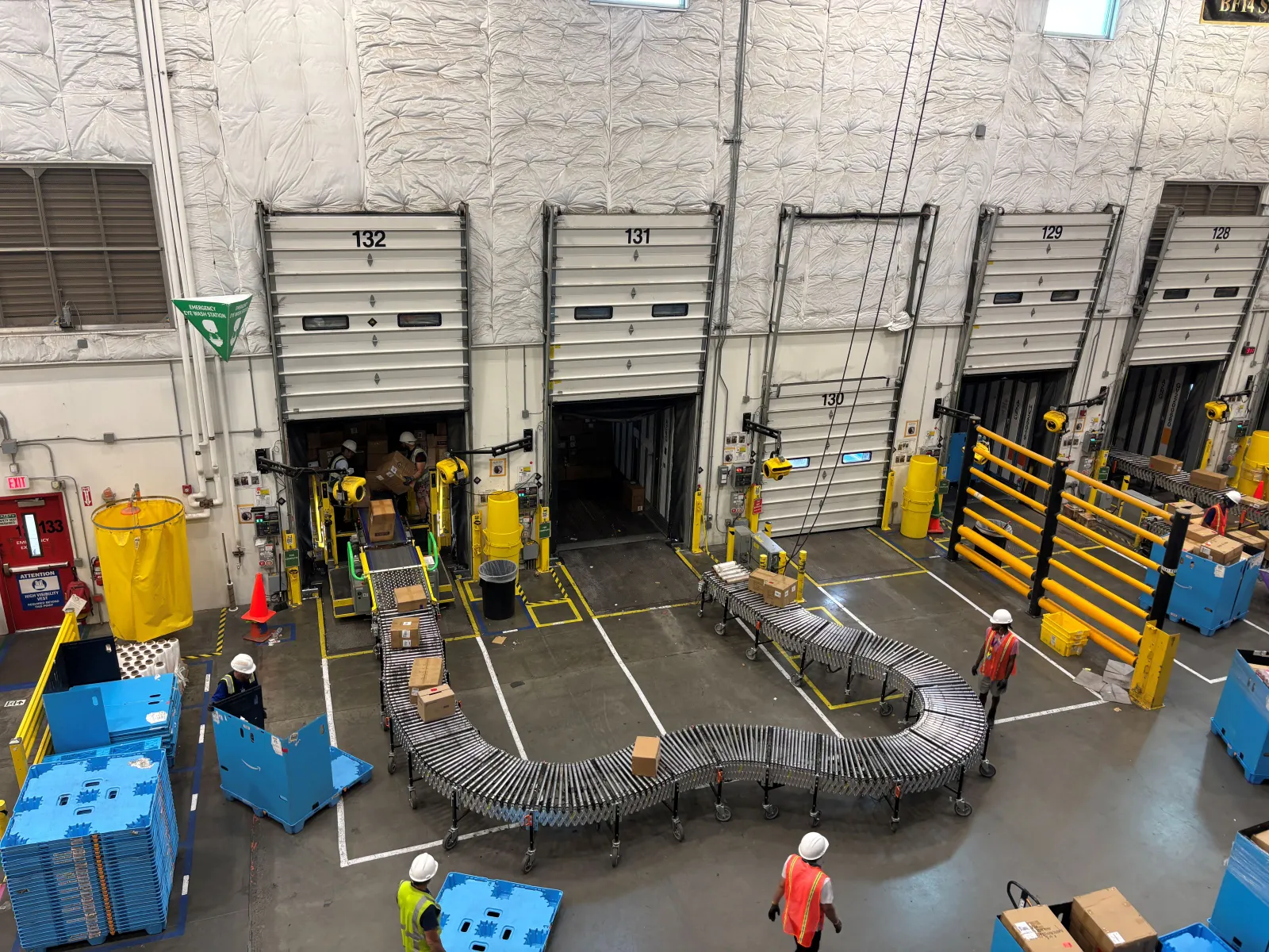 Boxes are being unloaded from a truck onto a conveyor belt with employees working in the background.