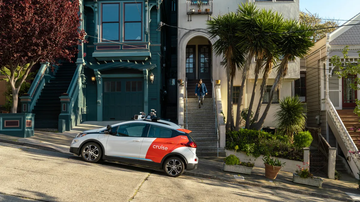 A small white car with a red rear door and a sign that reads "Cruise" is stopped on a steep street in San Francisco as a person walks down the steps of a home.