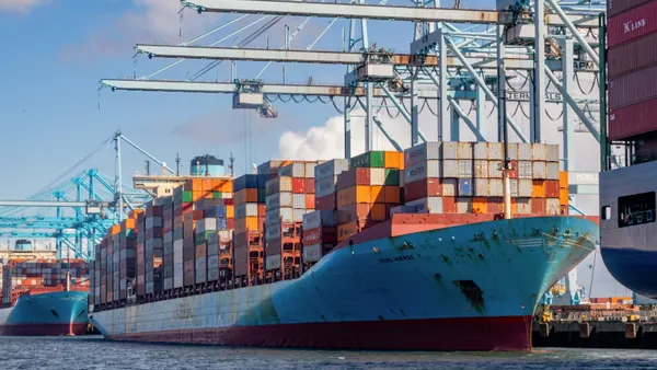Dozens of multi-colored containers are stacked on a blue cargo ship docked at a port under cranes. The cargo ship is between two other vessels.