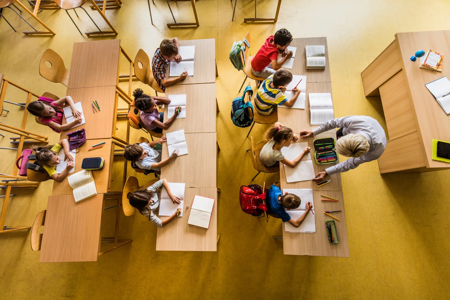 A photo from above-looking-down of an elementary school classroom with rows of desks. Students are stilling at the desks with notebooks in front of them. An adult is standing at the front of the class