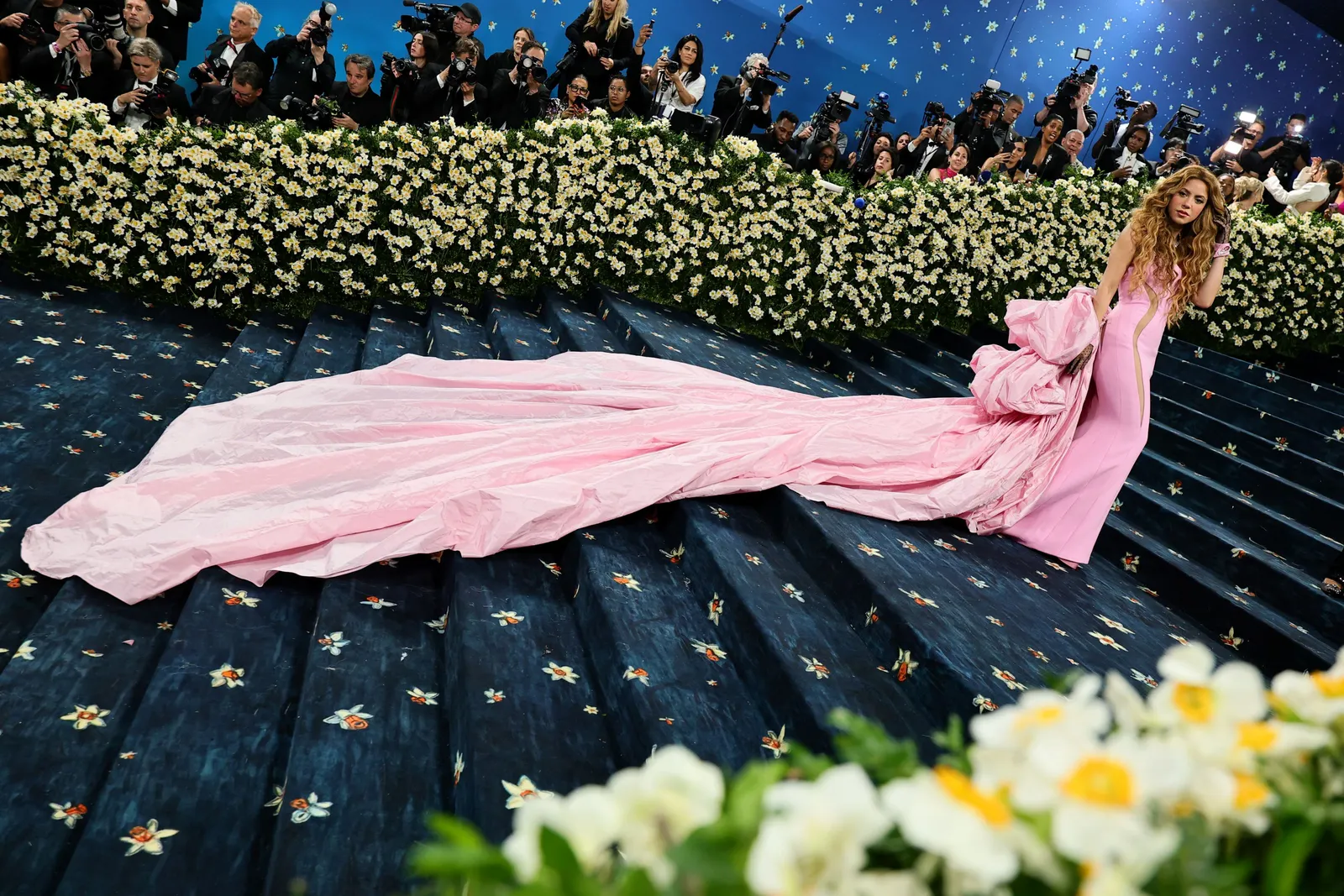 A woman in a voluminous pink strapless dress poses on a blue carpet.