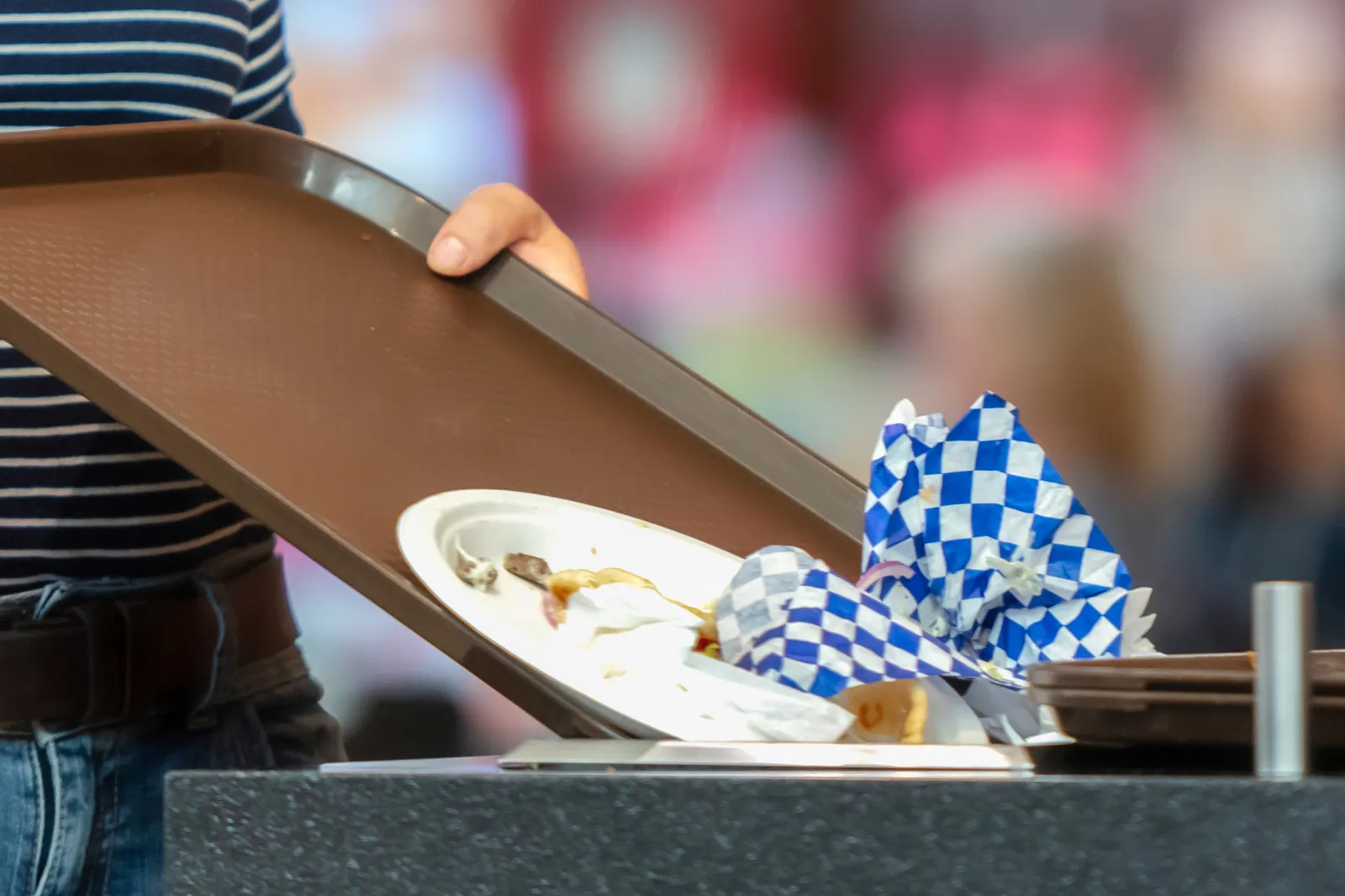 Close-up of a woman with a food tray, tossing her white plate and hamburger wrapper into the trash.