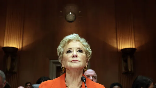 A person is photographed from the front and below. Only the person's head, neck and shoulders can be seen. They are in a room with wood paneling. Others are seated behind the person.