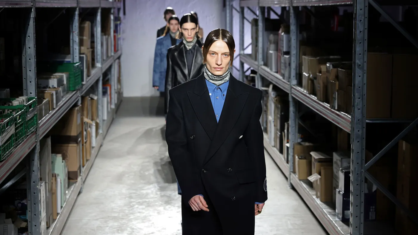 Models walk in a line between metallic warehouse shelves lined with boxes.
