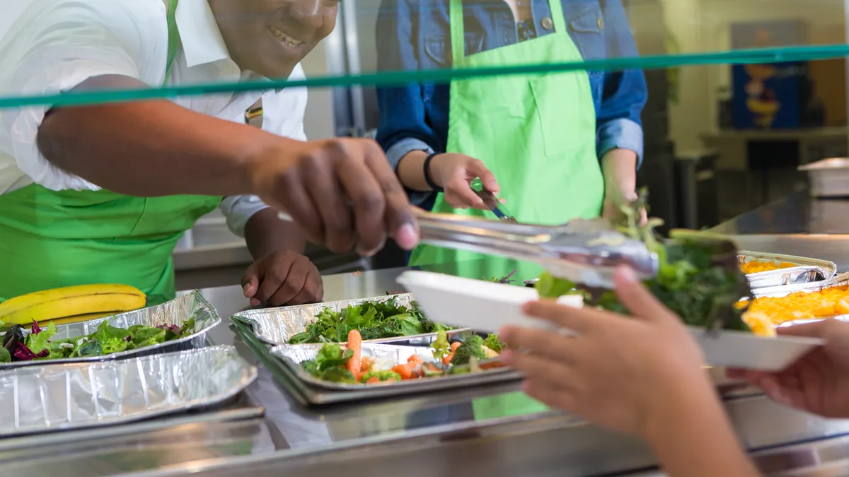 A school cafeteria worker serves a student food on their tray while waiting in line.