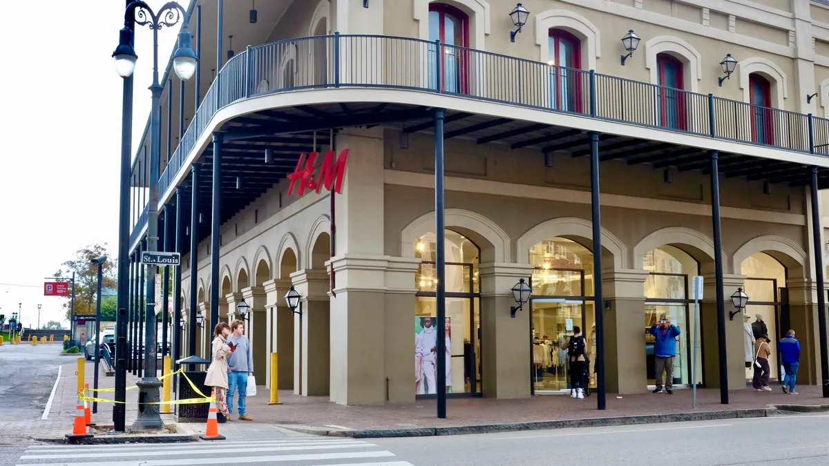 A street lamp and a couple of people to the left of a large corner store with a red "H&M" sign, as others stroll and window shop.