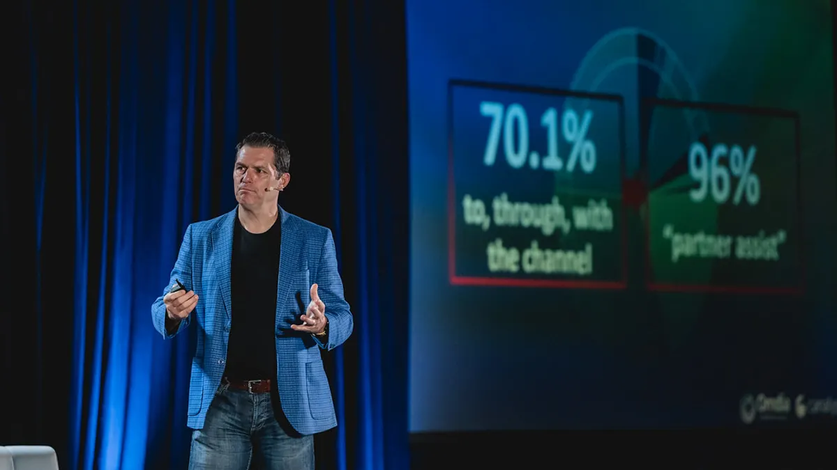 A man in blue jeans and a blue blazer gives keynote speech with a white chair and a large monitor in the background.