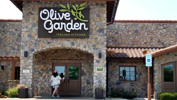 An iamge of a stoneworked building with a white logo that says Olive Garden. A woman wearing a white shirt and red bandana is at the door about to enter the restaurant.