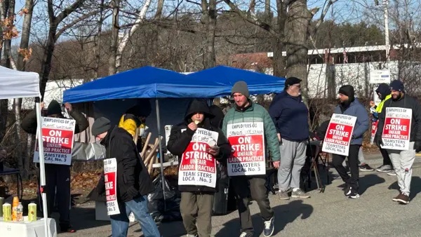 Multiple people gathered in front of an industrial facility hold picket signs saying they are part of the Teamsters Local 3 and are on strike.