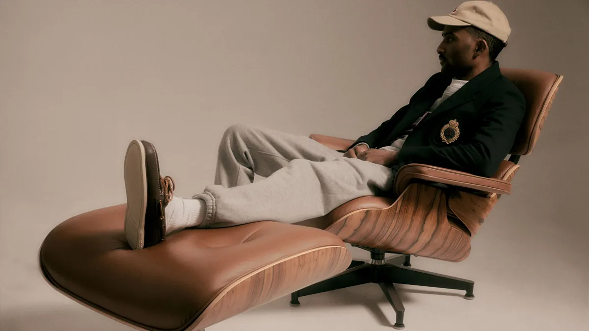 A person wearing a ballcap, cardigan, sweatpants and boat shoes lounges on a brown leather Eames-style chair.