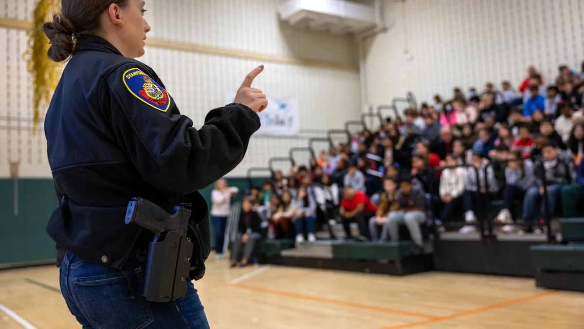 Students sit in rows in bleachers in a school gym listening to a police officer who is standing on the gym court floor.