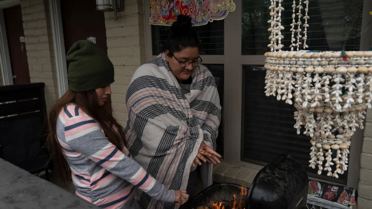 Two people stand next to flames in a barbecue with their hands stretched out over the fire. One person is wrapped in a blanket.