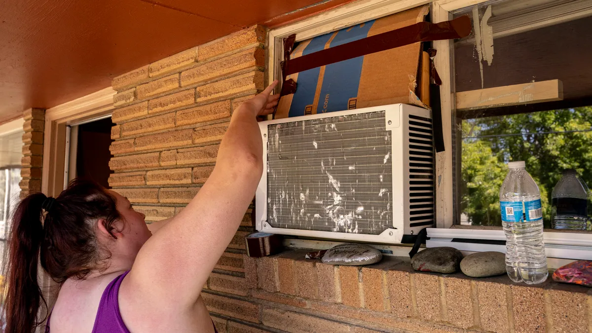 A person wearing a ponytail and purple tank top raches up to touch cardboard around a window AC unit on the outside of a building. A plastic water bottle sits on the windowsill.