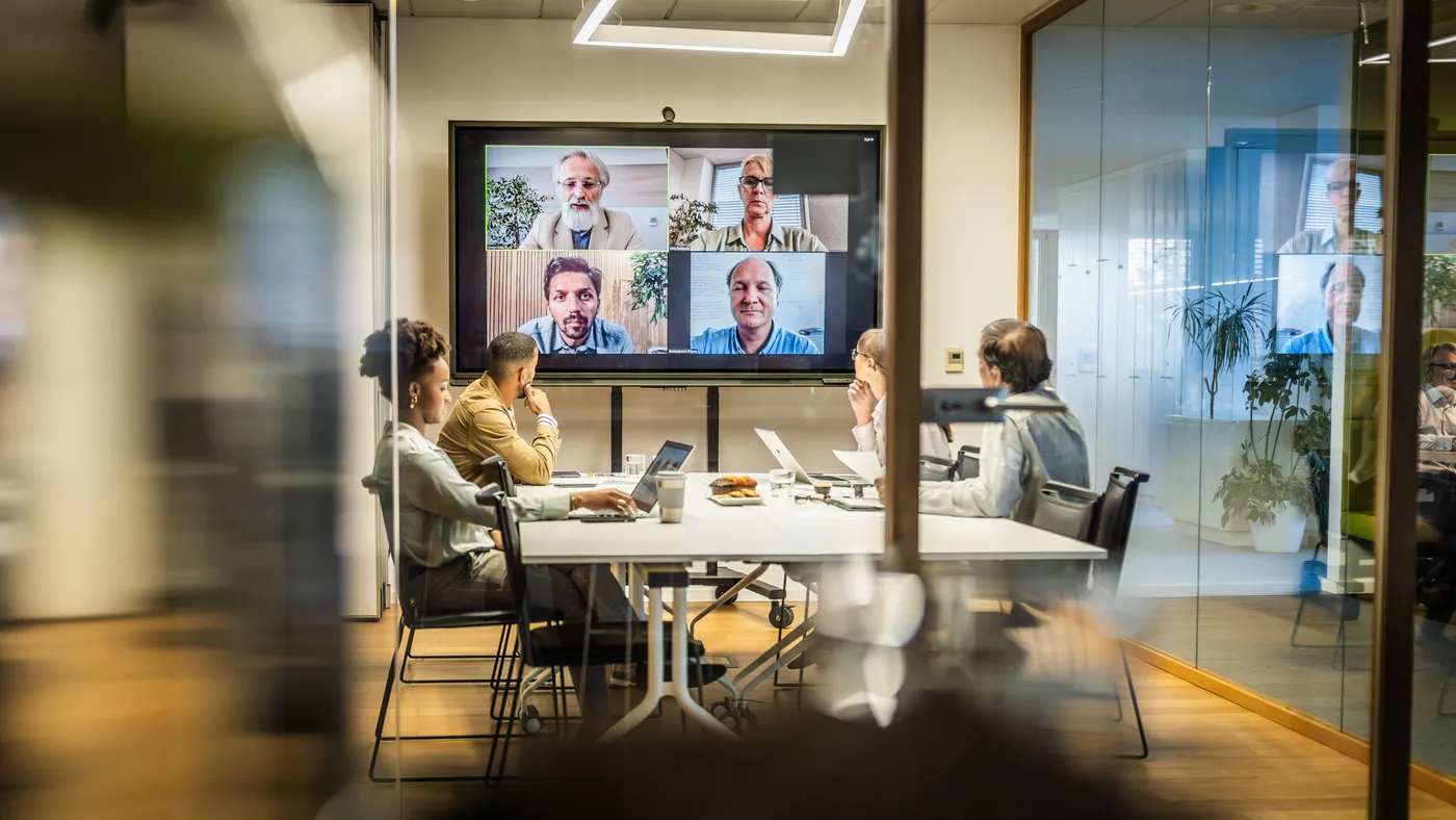 A group of employees attend a hybrid attendance presentation from a small meeting room.