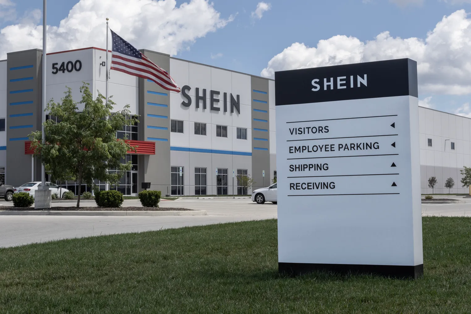 A closeup of a Shein sign directing people around its distribution center. The Shein building is seen in the background, with an American flag.