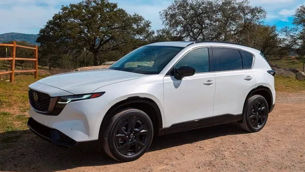 A white 2026 Mazda CX5 crossover parked outdoors with a wooden fence, trees and hills in the background.